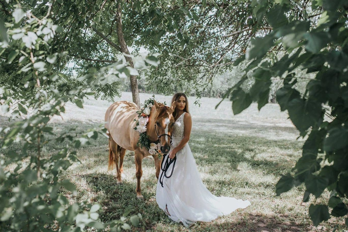 A bride in a boho wedding dress leading a horse through a wooded area. 