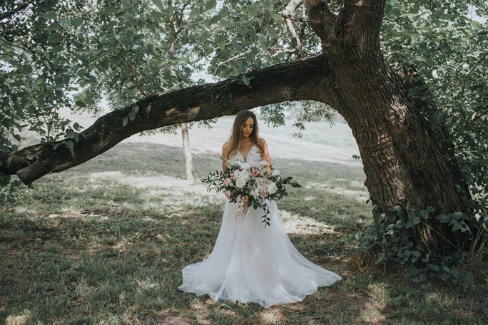 Bride wearing a boho wedding dress holding a large bouquet under a tree. 