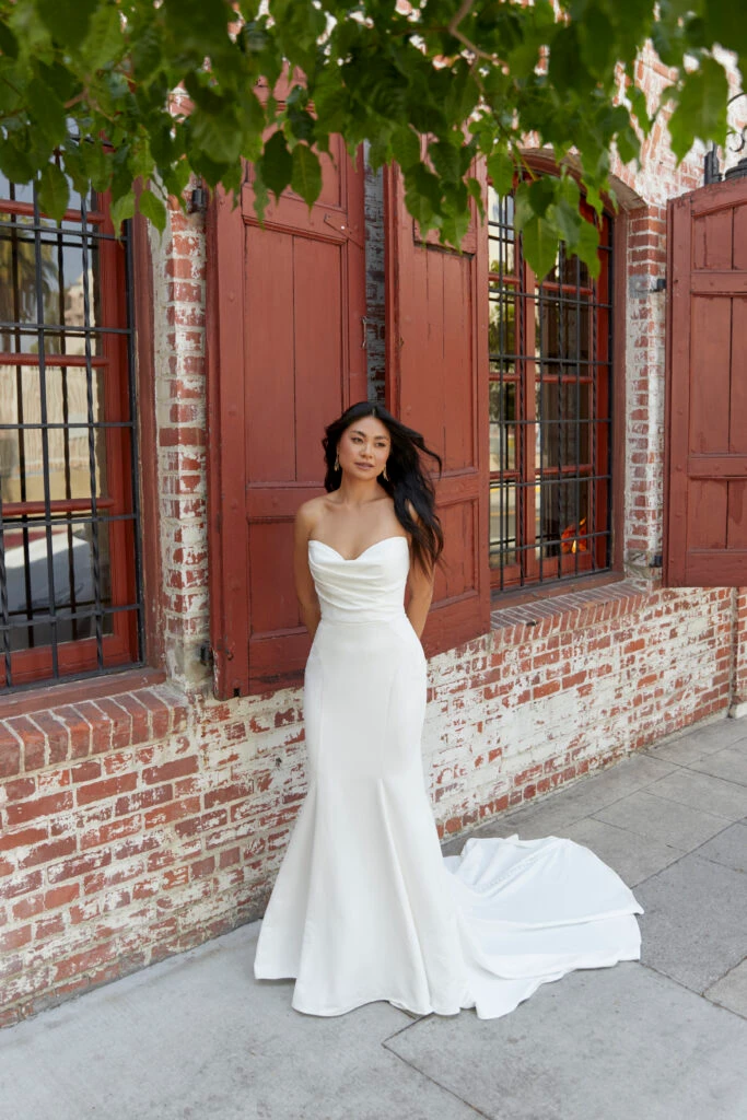 Bride in a strapless wedding dress with a sweetheart neckline, standing against a rustic brick wall with red shutters, framed by green leaves above.