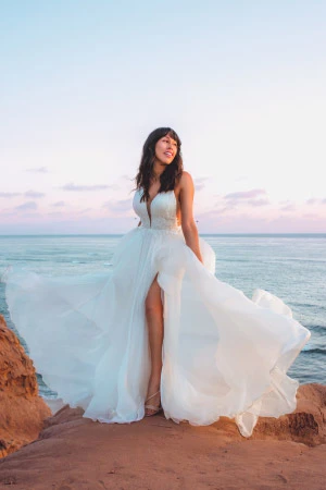 Asian woman with long brown hair standing on a beach wearing white wedding dress with a flowing chiffon skirt and a slit.