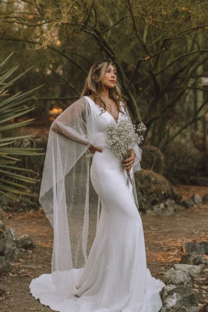 Woman with long brown hair wearing a white boho-style wedding dress with a sheer cape and v-neckline.