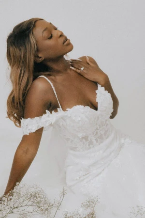 African-American woman wearing a white wedding dress with lace details, thin straps and off-the-shoulder sleeves.