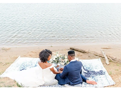 Bride sitting on a rug on the beach with her husband.