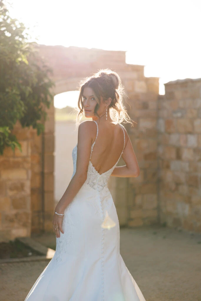 Braut in einem tief ausgeschnittenen Hochzeitskleid mit zarten Trägern, steht im Sonnenlicht nahe einer Steinmauer und blickt sanft über die Schulter.