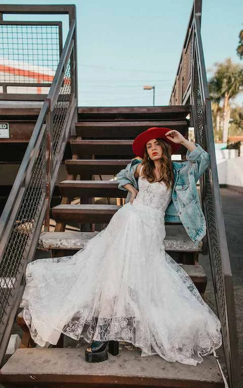 Bride wearing boho lace wedding dress and red wide-brimmed bridal hat sits on stairs.