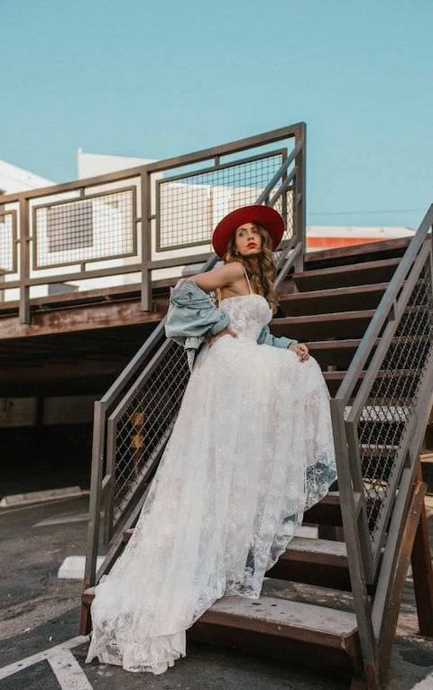 Bride wearing boho lace wedding dress and red wide-brimmed bridal hat stands on stairs.