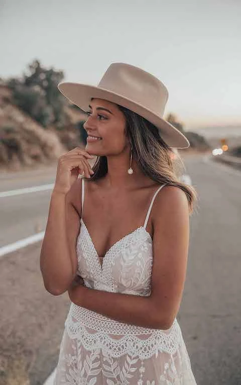Bride stands on road wearing lace boho wedding dress and wide-brimmed bridal hat.