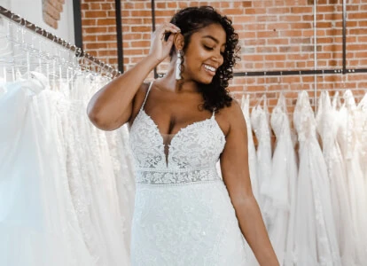 Woman of color wearing a sparkly lace, wedding dress, standing in front of a rack of wedding gowns at True Society by Belle Vogue Bridal.