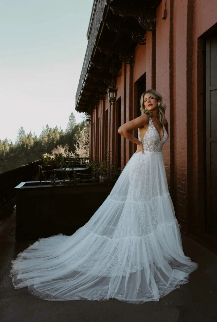 a bride is standing next to a barn in a tired a-line wedding dress with a floral lace bodice, cece by All Who Wander. She has hands on her hips and is looking to the left over her shoulder.