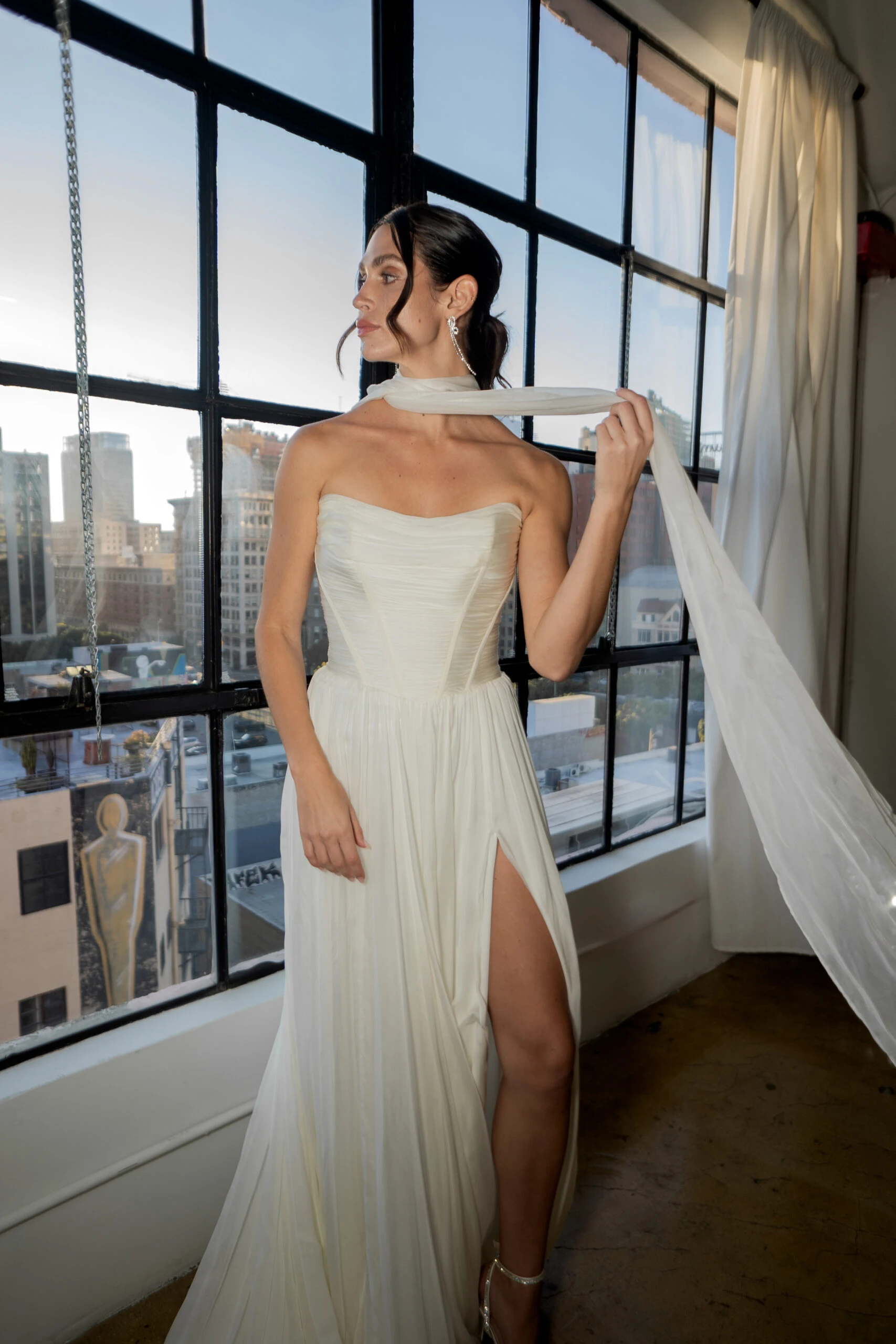 a bride is standing by a window in a downtown loft wearing a simple silk wedding dress with a matching neck scarf, Avalon by All Who Wander. She is facing the left and flipping the scarf over her neck.