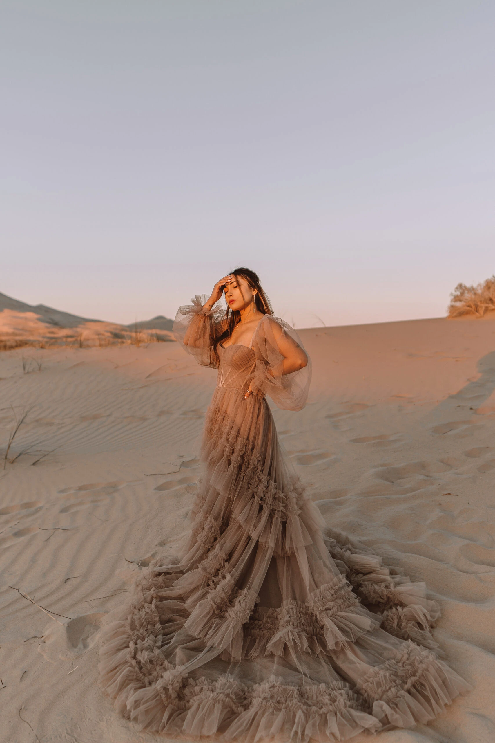 A bride is standing in a san dune in a light brown ruffled wedding dress, Sutton by All Who Wander. She has the train wrapped around her feet and has her hand up looking into the sunset.