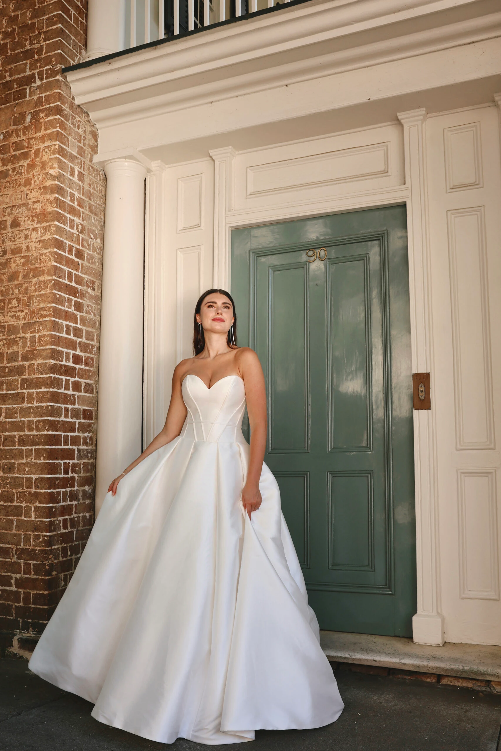 Braut trägt ein strukturiertes, trägerloses Ballkleid-Hochzeitskleid vor einem Brauthaus mit grüner Tür.