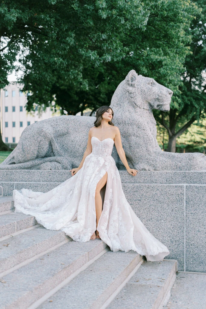 A bride is wearing a sparkly ballgown wedding dress, Martina Liana Luxe LE1231, in front of a lion statue. She is leaning back on her hands and showing off the slit of her dress. She is looking over her left shoulder.