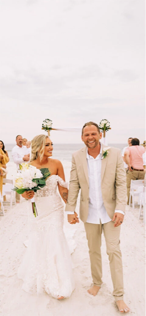 Real True Bride Danielle and her groom Kaleb smile as they walk hand in hand down a sandy beach aisle after their wedding ceremony.