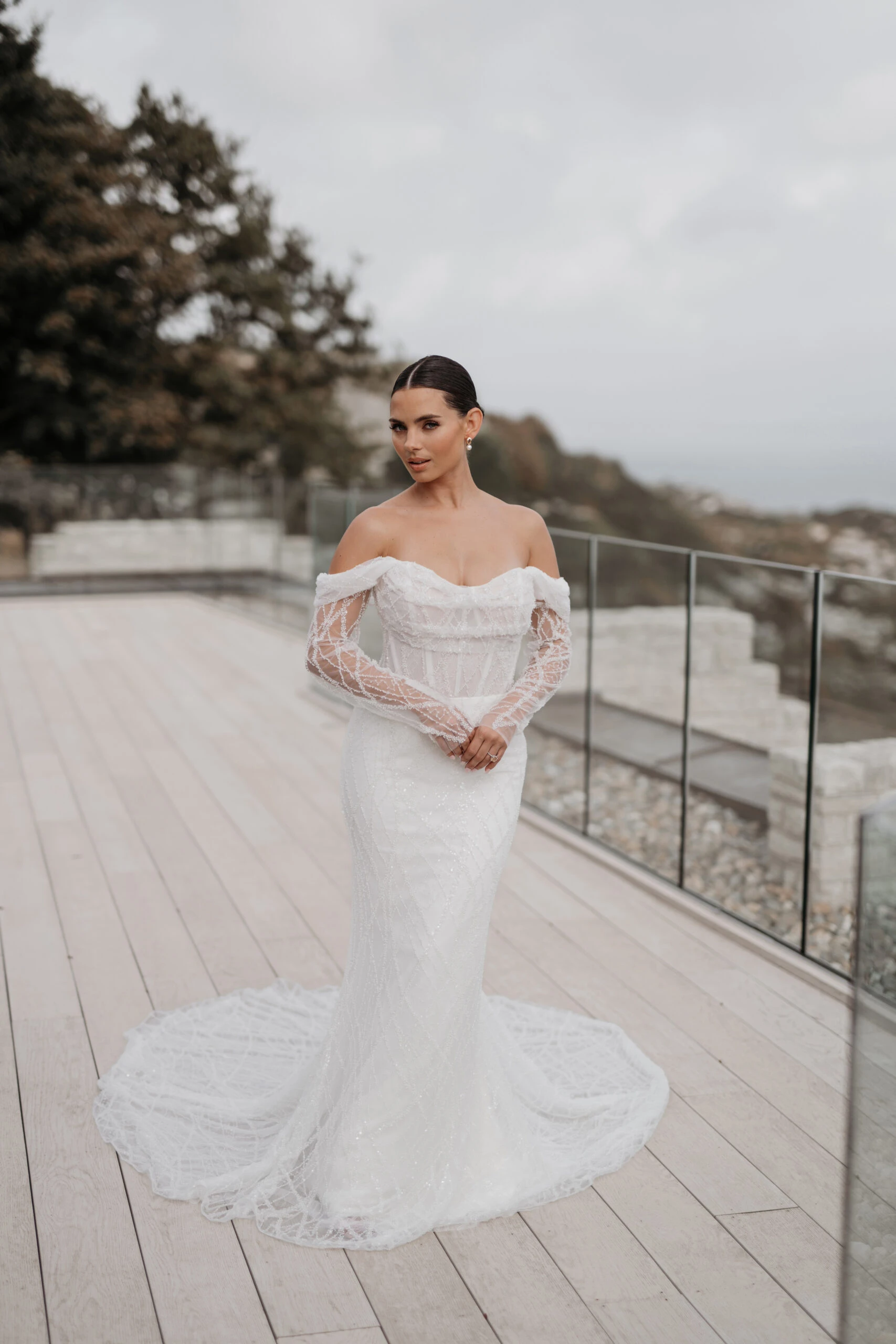 a bride is standing on a patio wearing a sexy lace mermaid wedding dress with off the shoulder long sleeves, LE1318 by Martina Liana Luxe. She has her hands clasped in front of her and is glancing up to the camera.