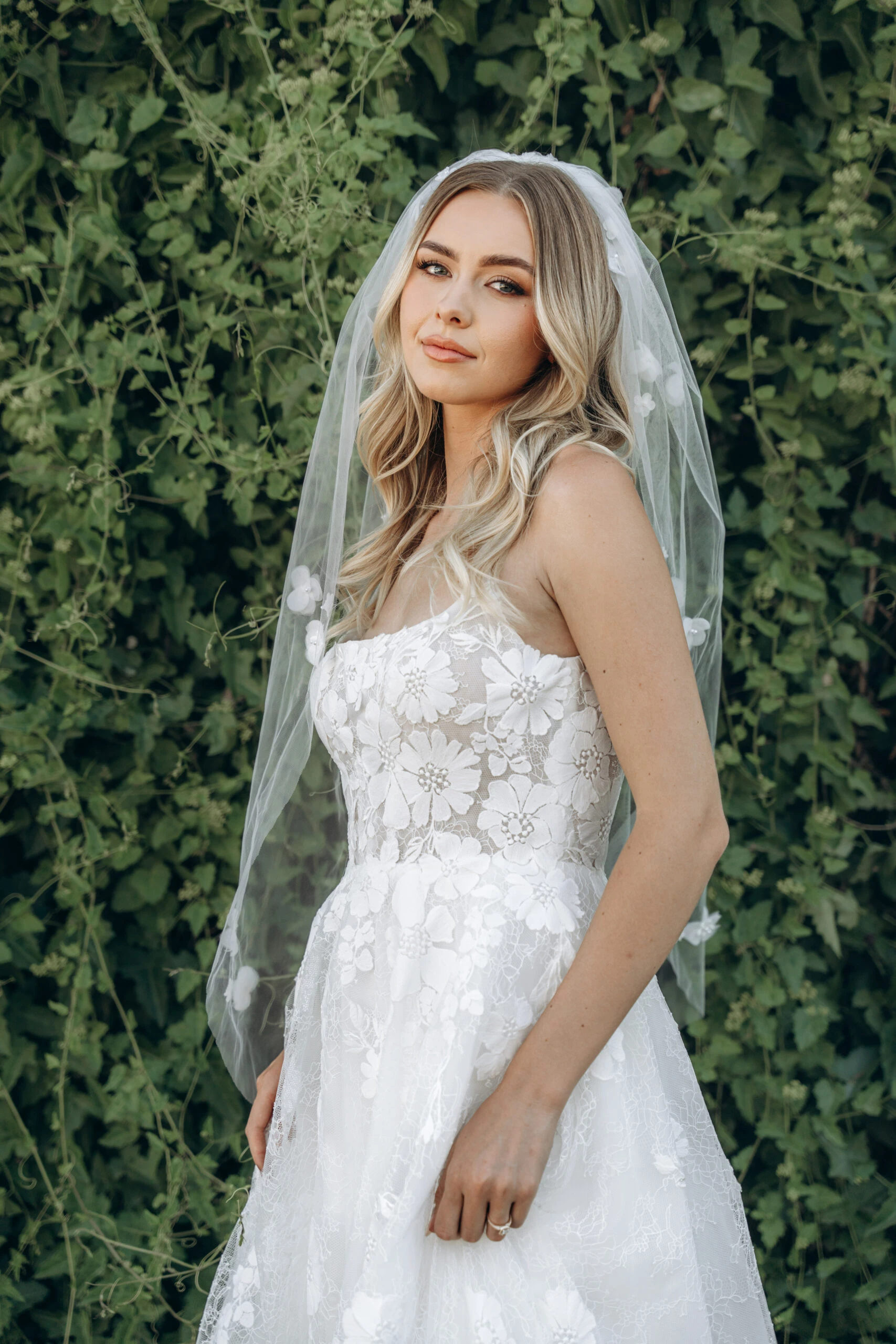 A bride stands against green foliage in a strapless floral lace wedding dress with a delicate veil, showcasing her romantic style with soft, flowing blonde waves.