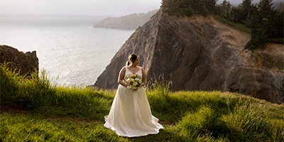 Plus-size bride wearing a simple ballgown wedding dress standing on grass hill with ocean view.