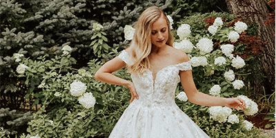 Bride wearing an off-the-shoulder lace A-line wedding dress stands in front of white floral bush.