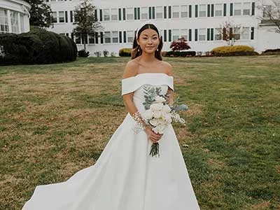 Bride holds white bouquet wearing designer simple off-the-shoulder ballgown wedding dress.