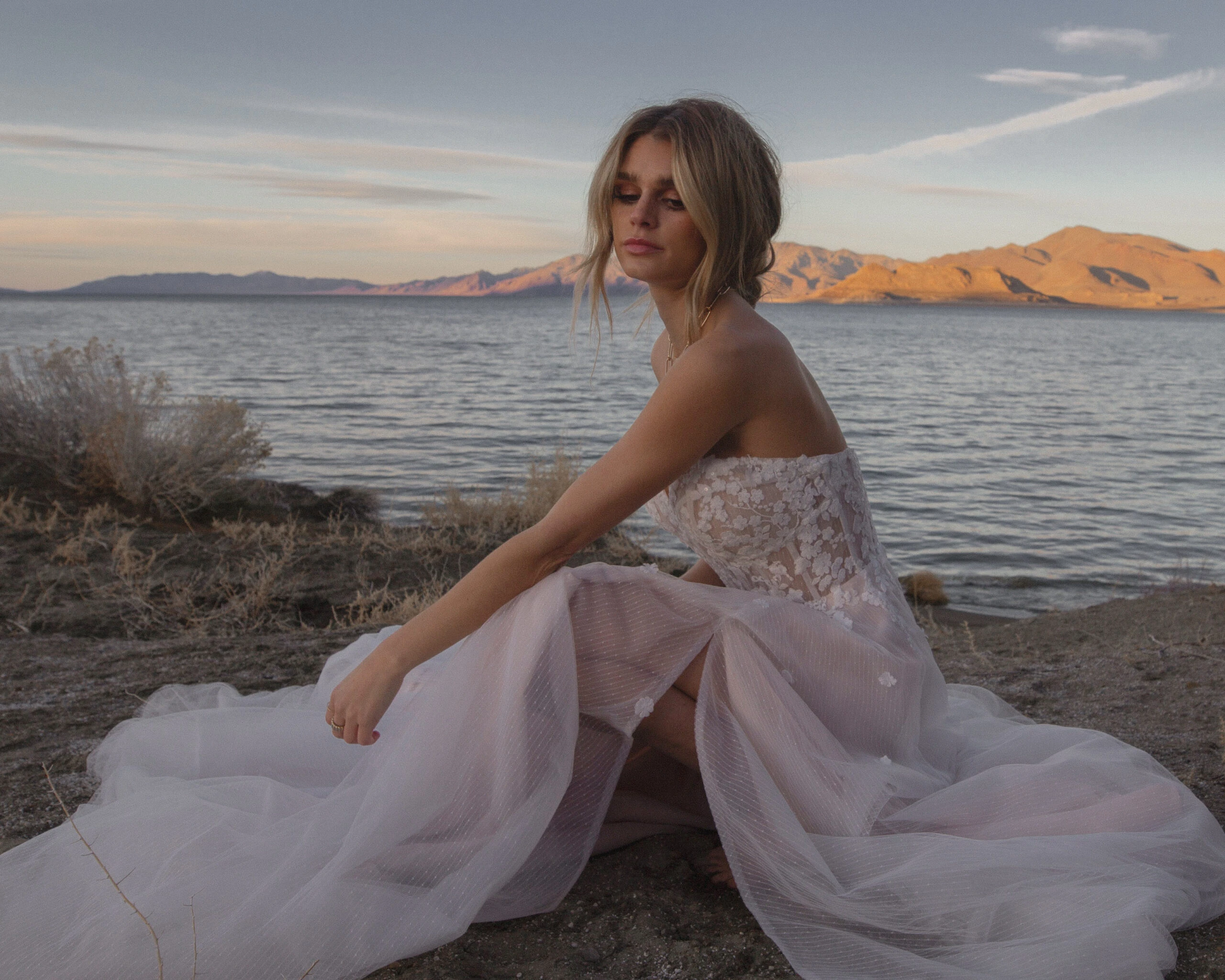 Bride wearing floral lace and tulle strapless wedding dress sitting on beach sand.