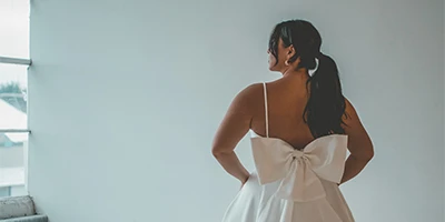 Bride wearing an open back wedding dress with back bow detail faces a wall.