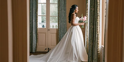 Bride wearing simple strapless ballgown wedding dress faces a window.