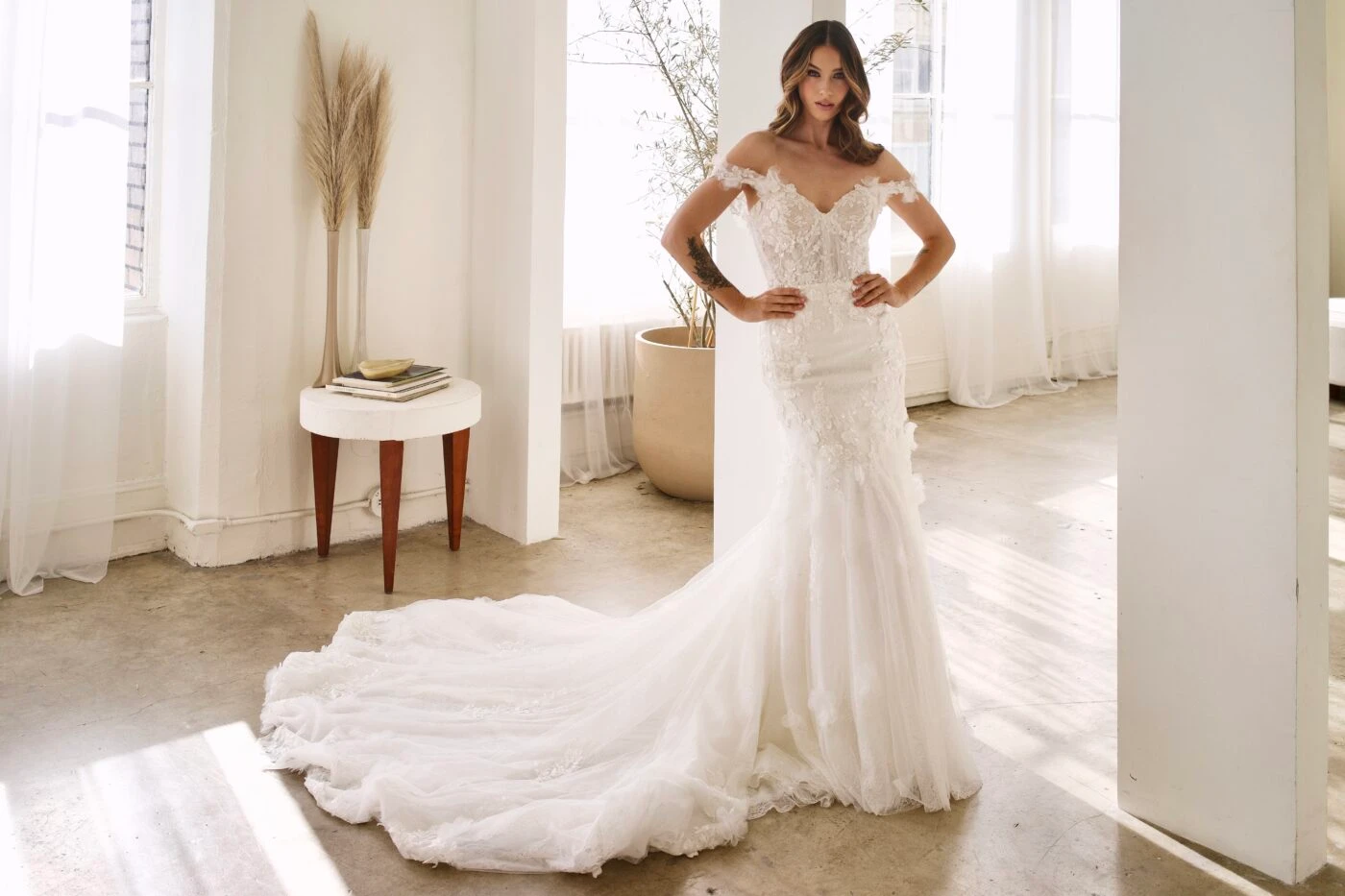 bride is standing in a well lit room with large windows wearing a lace column wedding dress with off the shoulder straps - Martina Liana Style 1612. She has hr hands on her hips and is facing the camera.