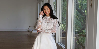 Bride wearing lace long sleeve wedding dress stands in front of large windows.