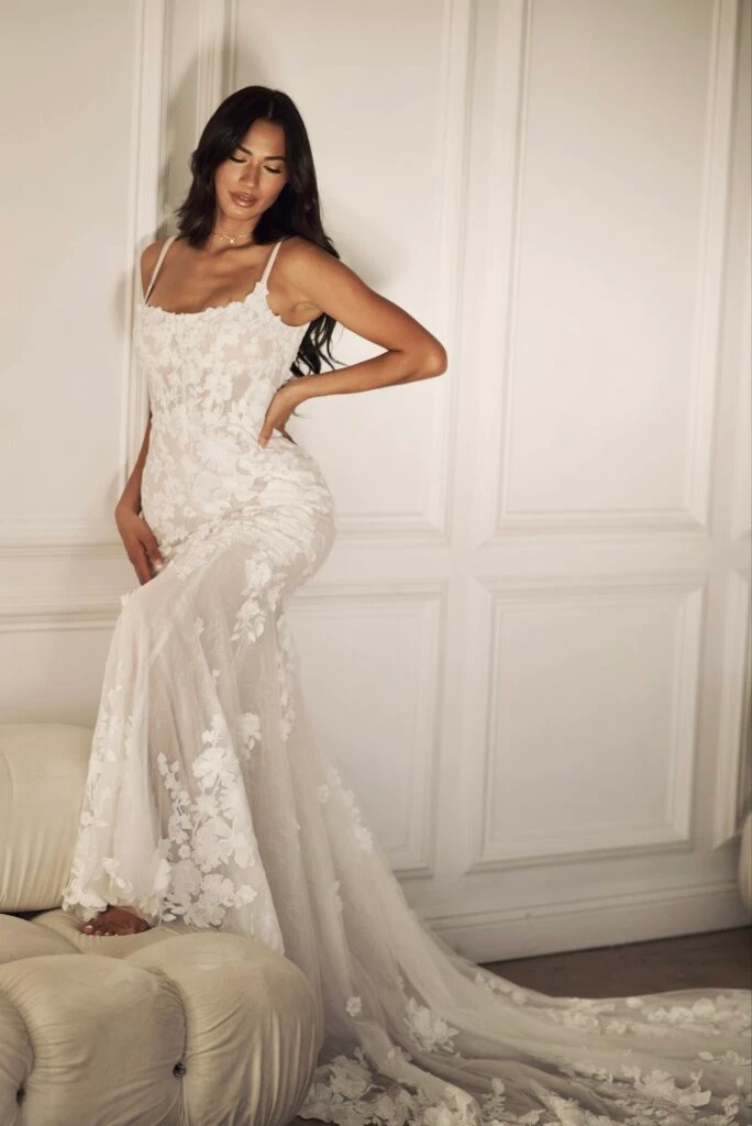 A bride is standing with her hand on her hip and glancing downward. She is wearing a lace mermaid wedding dress with a scoop neckline and spaghetti straps - Martina Liana Style 1767.