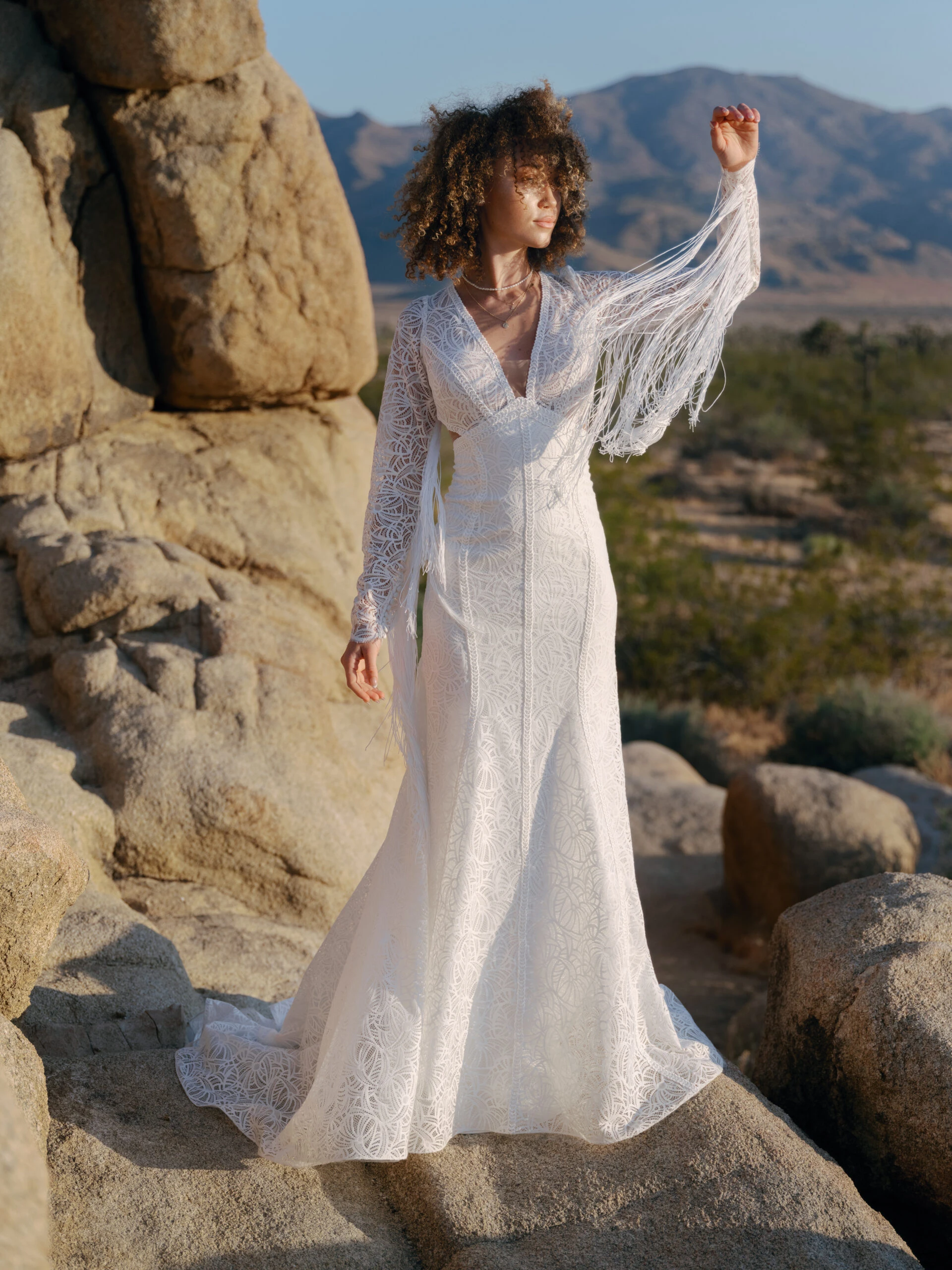 a country bride is standing in the desert in front of a rock looking into the sunset. She is wearing a long sleeve wedding dress with tassles and a keyhole back, Cash by All Who Wander