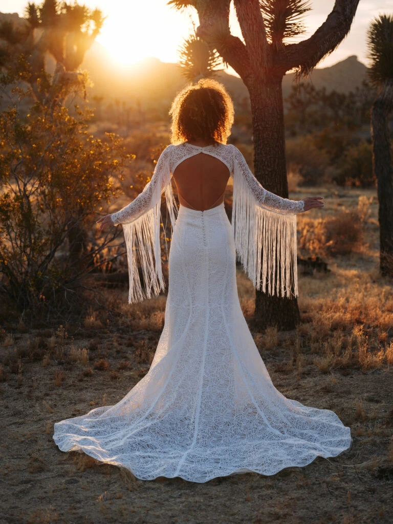 a bride is standing with her back to the camera in front of a cactus. She is wearing Cash by All Who Wander, a boho long sleeve wedding dress with keyhole back and tassel details.