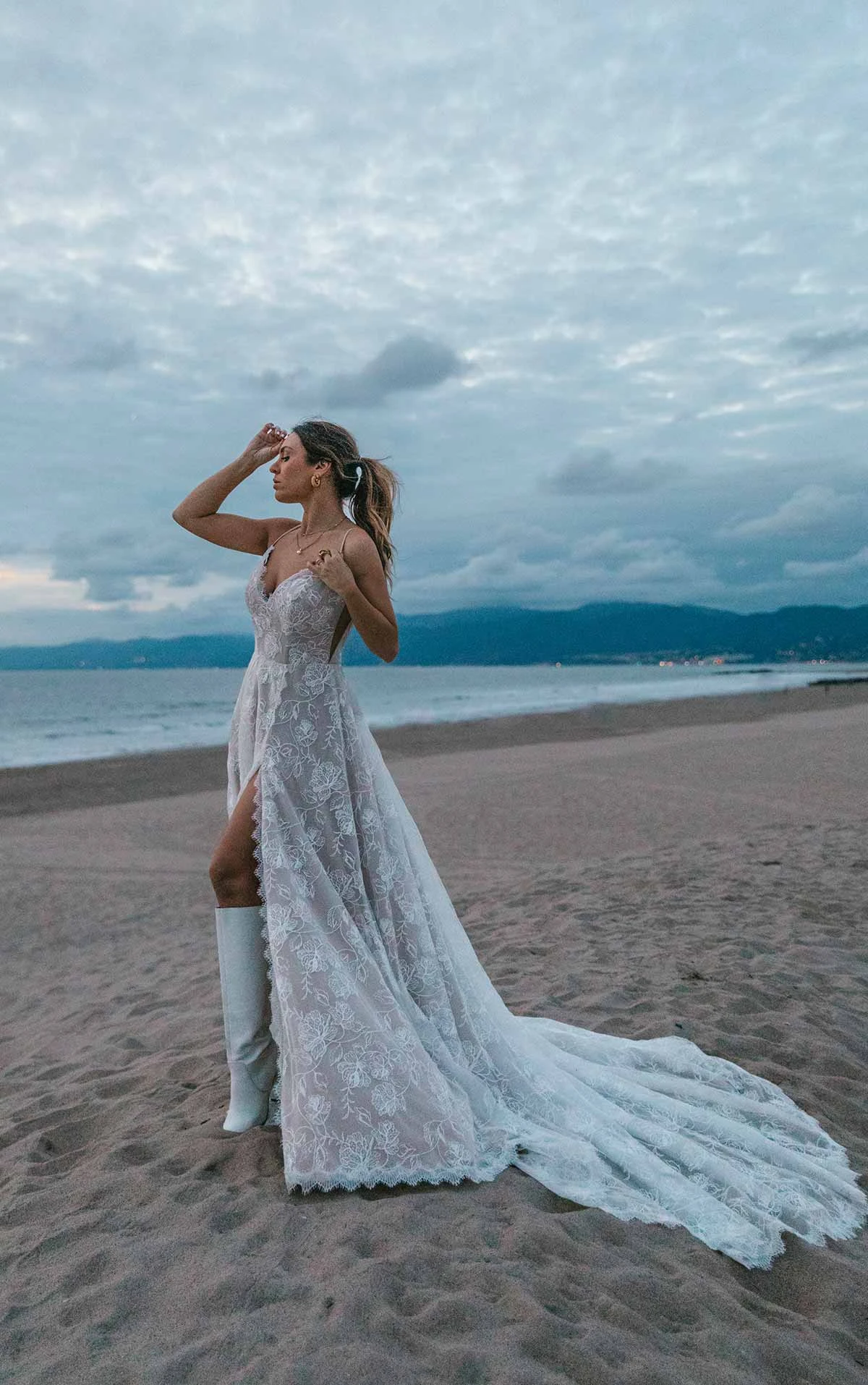 bride is standing on a beach wearing luna by All Who Wander, a lace a-line wedding dress with a high slit and spaghetti straps. She is facing the left with one hand at her face shielding from the wind.