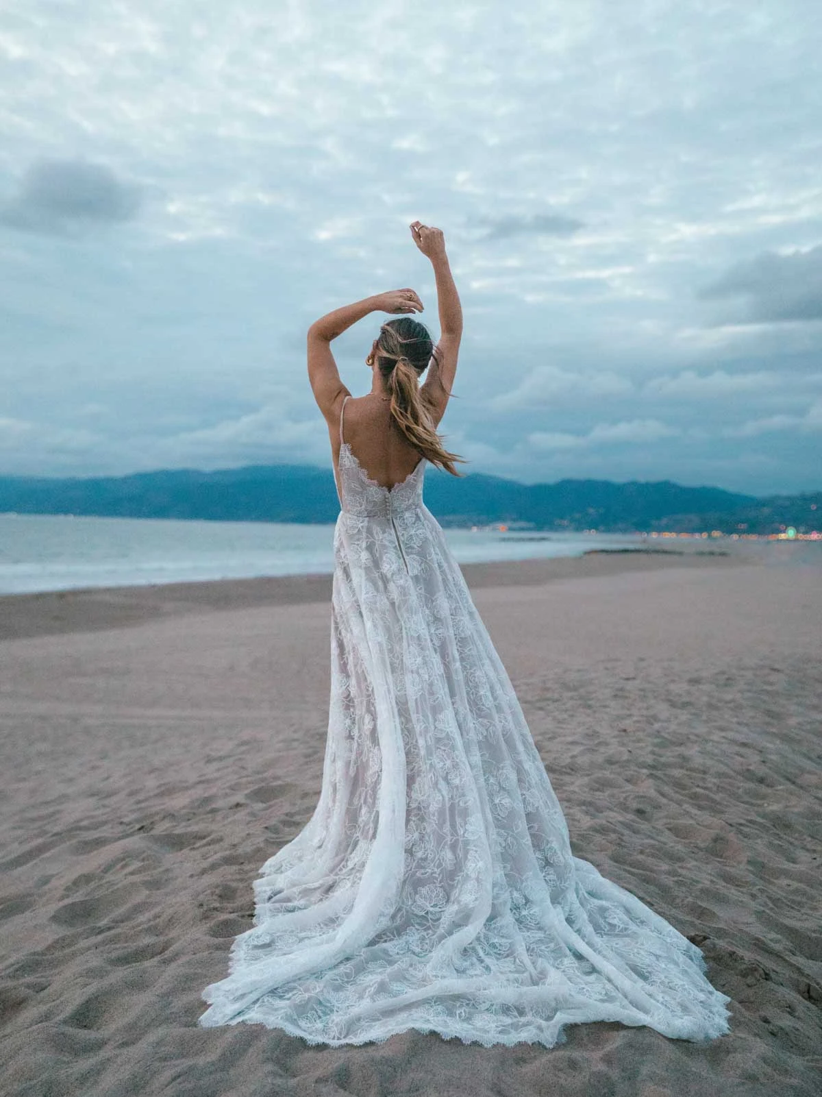 bride is standing on the beach in a flowy lace wedding dress, luna by All Who Wander. She is facing the waves with her hands above her head showing off the train and open back.