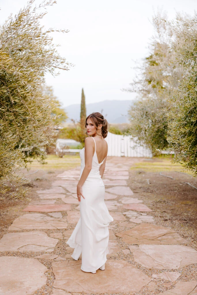 a bride is walking away on a stone and sand path looking over her shoulder. She is wearing a simple sheath wedding dress with a scoop back and spaghetti straps.