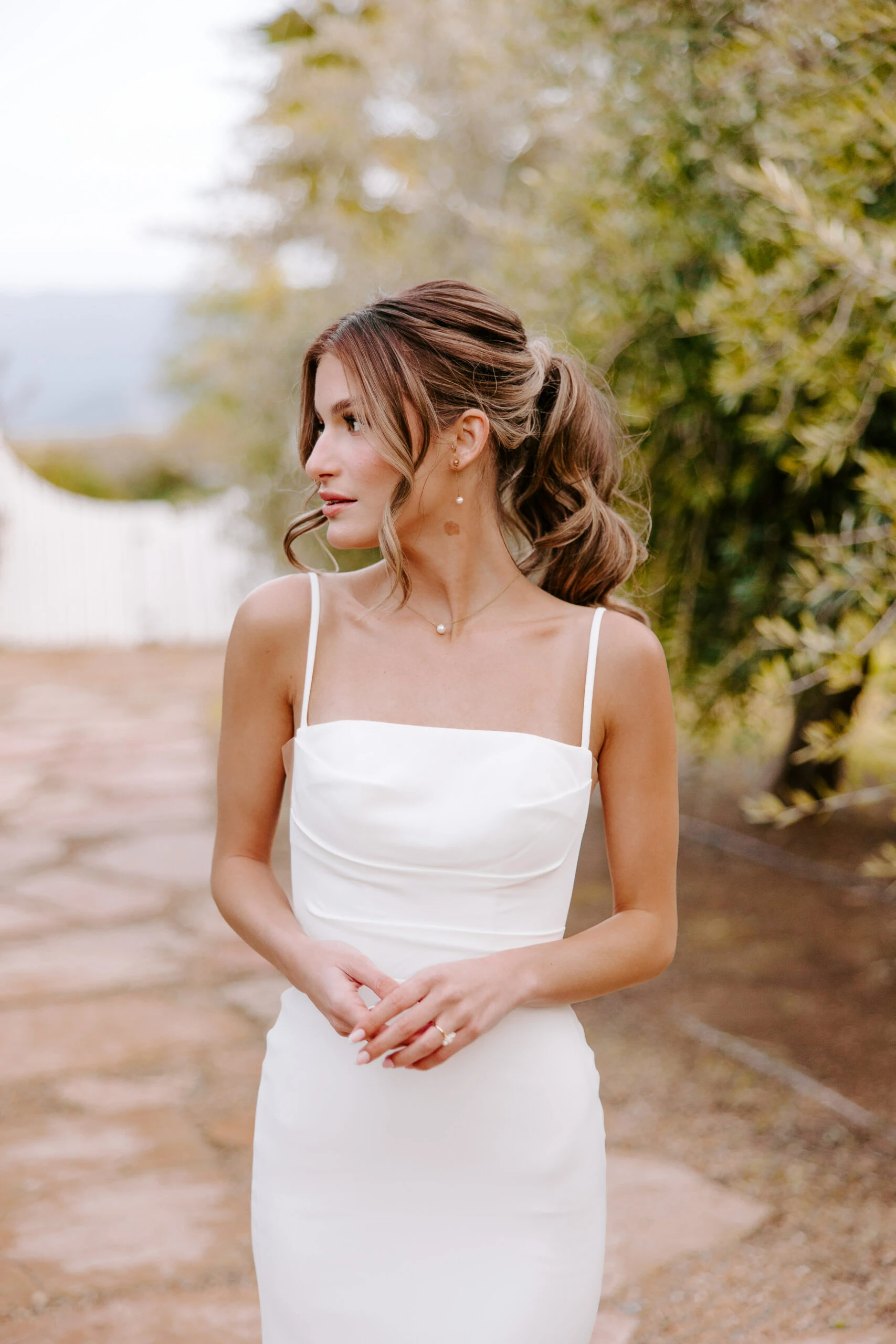 bride standing on a stone path wearing a simple sheath wedding dress with spaghetti straps, Jules by All Who Wander. She is looking to the left with her hands clasped in front of her.