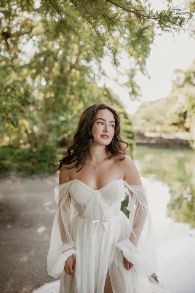 Bride standing near a river wearing a long sleeve off-the-shoulder dress