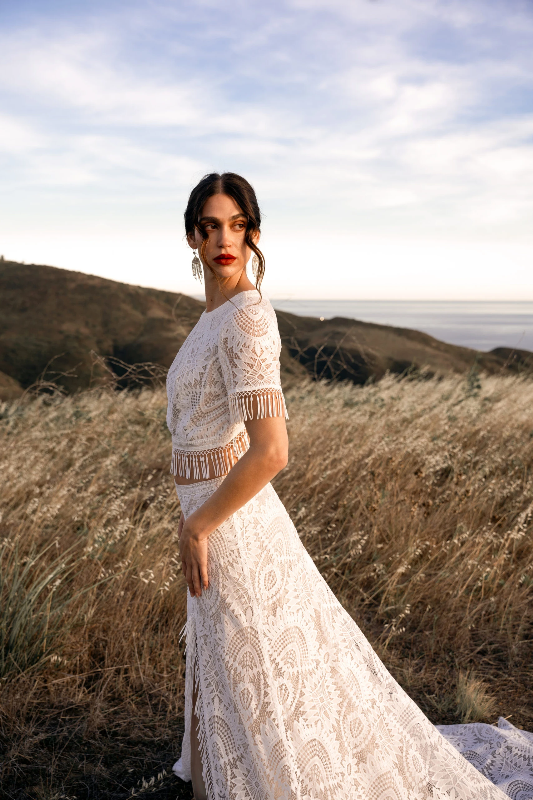 bride is standing in a field wearing a boho two piece wedding dress, Devyn by All Who Wander. She is looking over her shoulder with her hands on her hips, looking away from the camera.