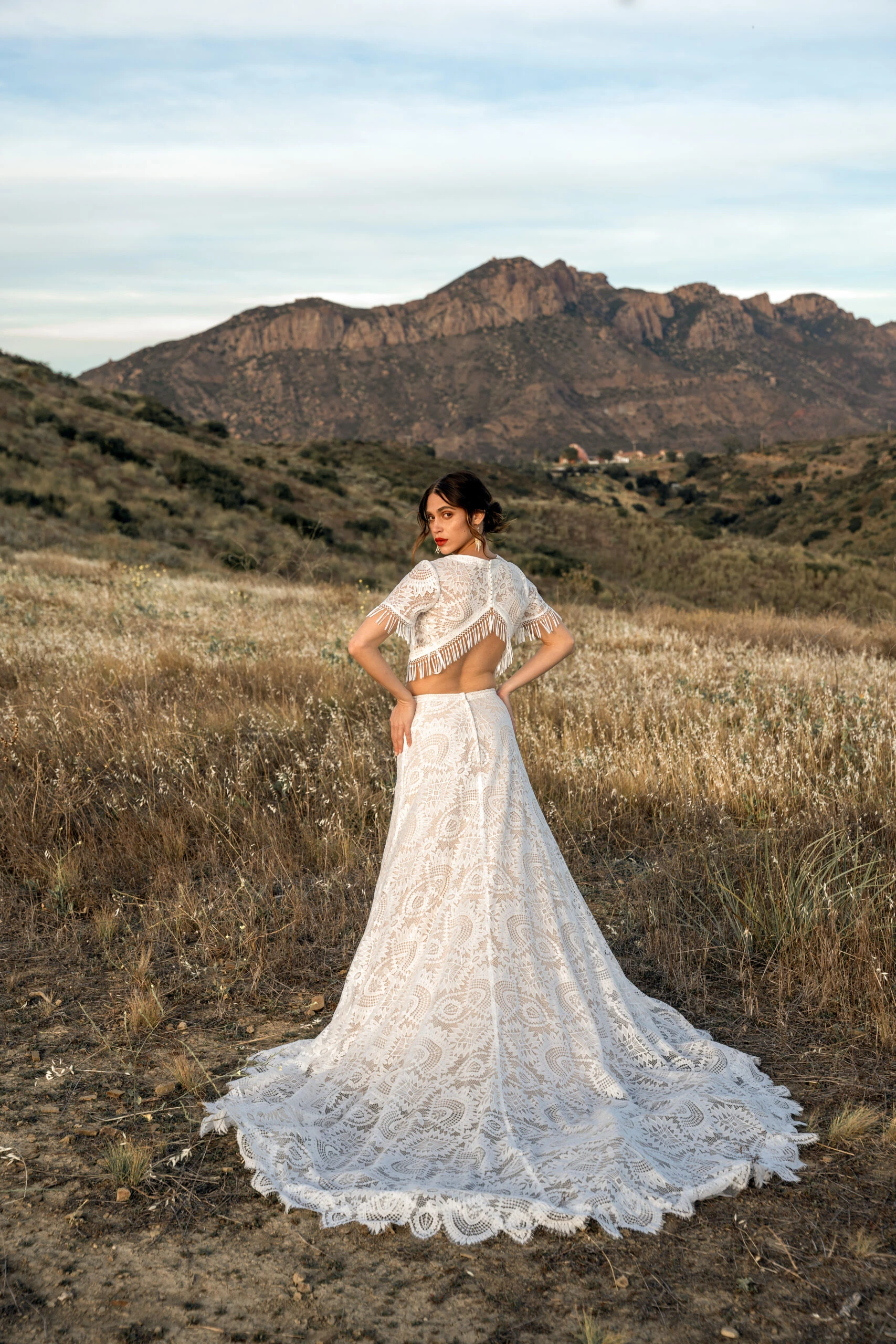 bride is standing in a large field with a view of mountains. She is looking back over her shoulders wearing a lace boho wedding dress with a back cutout, Devyn by All Who Wander.