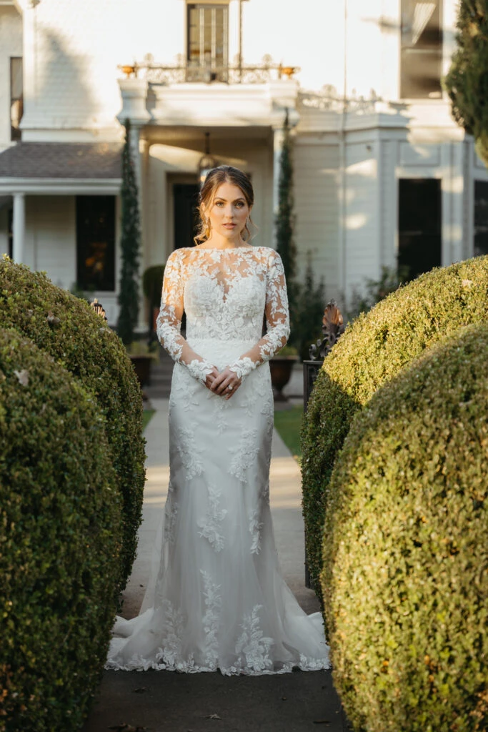 Bride wearing long sleeve lace wedding dress standing between bushes