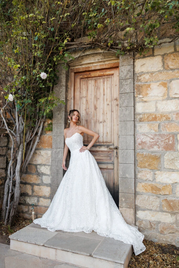 Braut trägt ein trägerloses Ballkleid-Hochzeitskleid aus Spitze und posiert vor einer rustikalen Holztür.