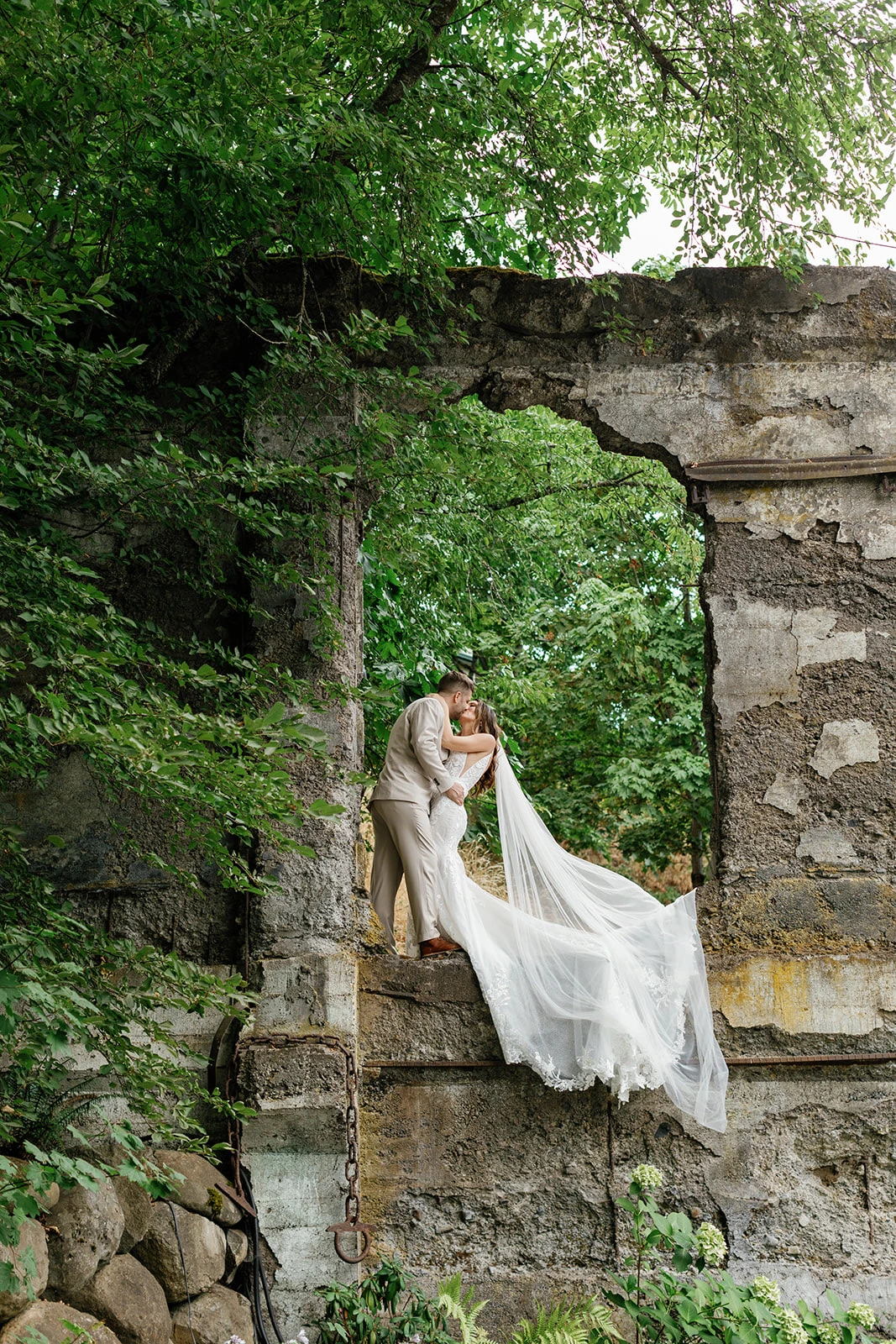 Bride and groom sharing a kiss on a rustic stone structure surrounded by lush green foliage, with the bride's long veil flowing down