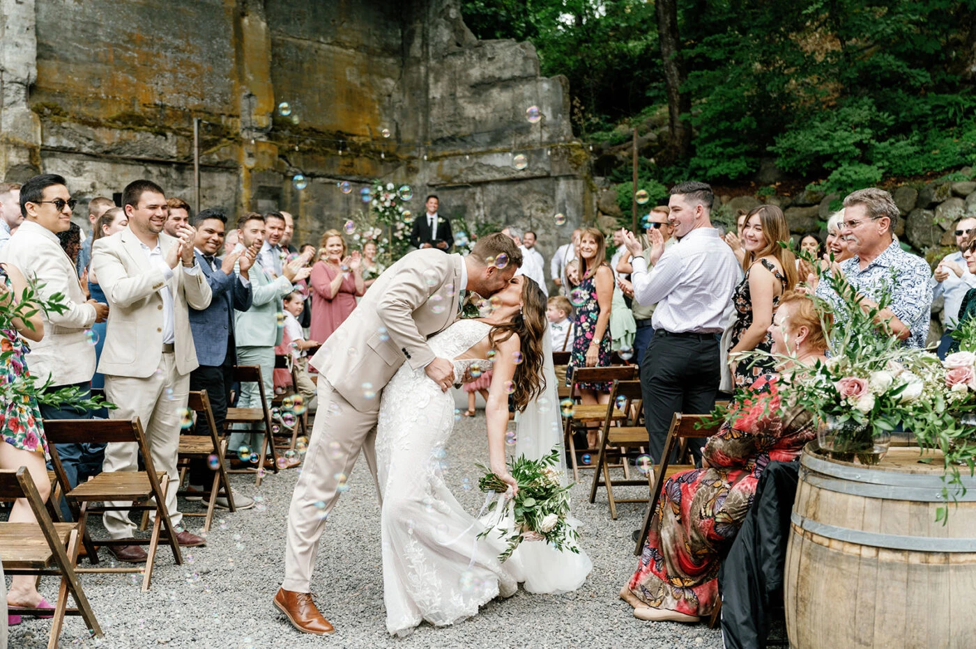 Bride and groom sharing a kiss while surrounded by cheering guests in an outdoor ceremony, with bubbles floating in the air