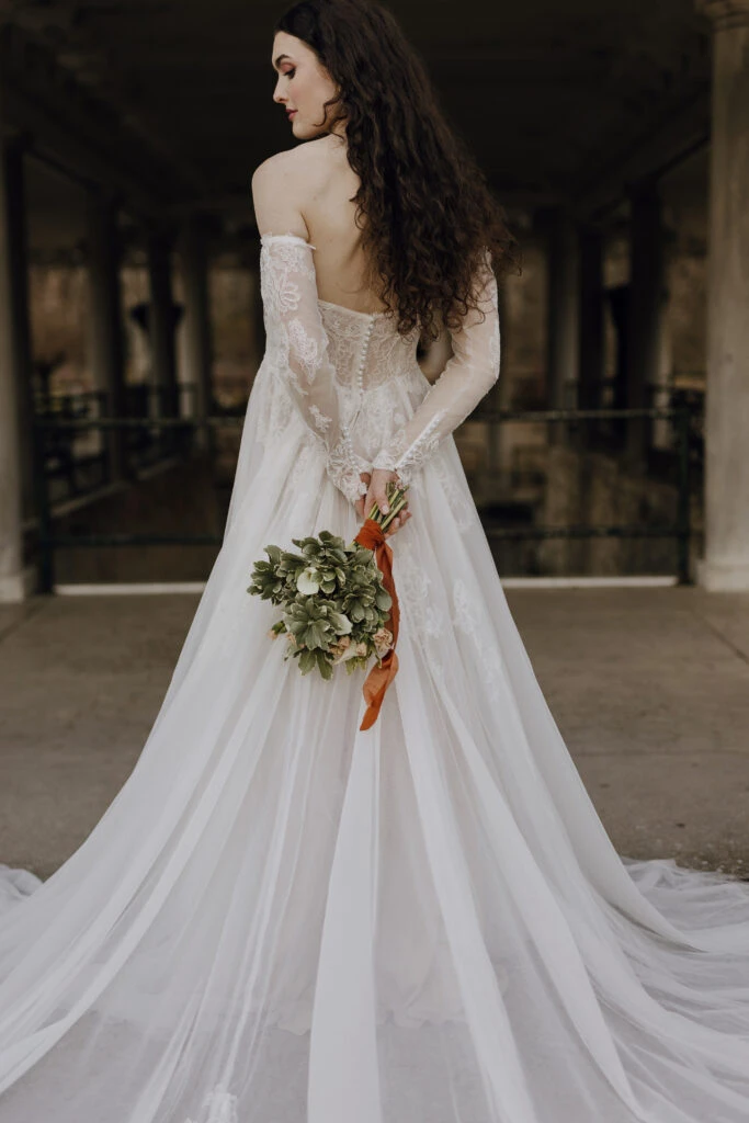 Back of bride wearing a long sleeve off-the-shoulder wedding dress while holding a bouquet of flowers