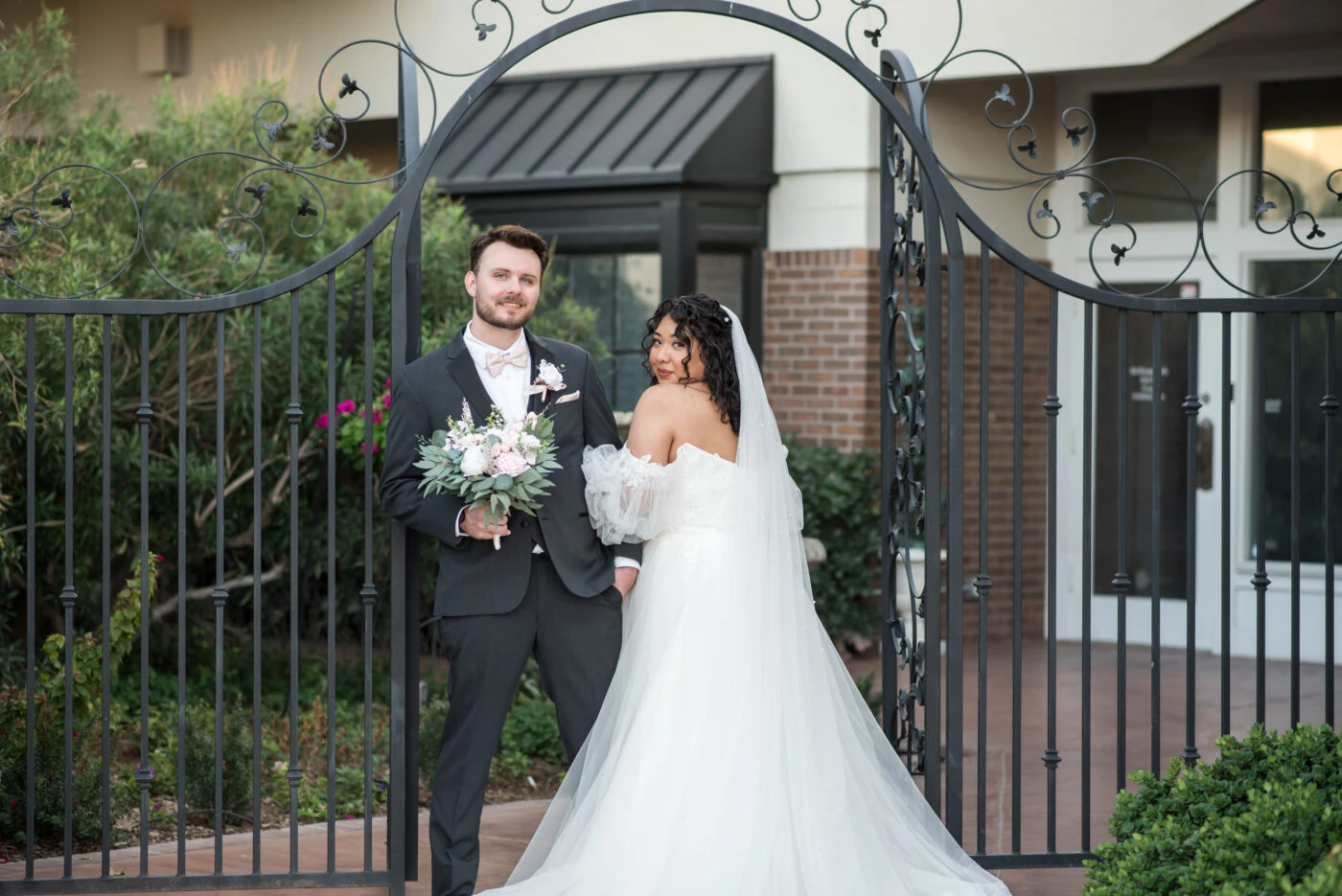 The bride and groom, holding a bouquet, smile while posing under a gate in a garden.