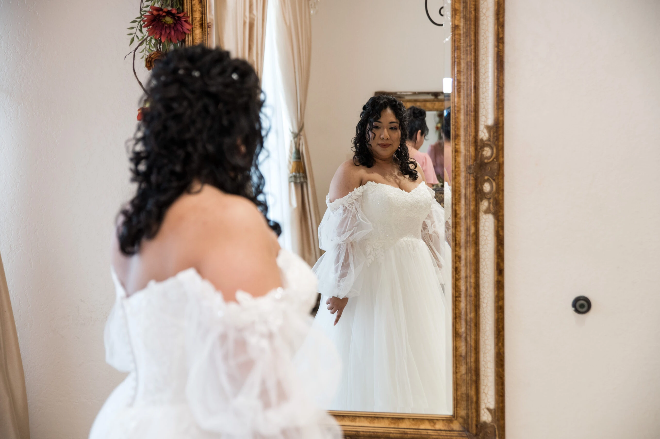 A bride with an off-the-shoulder wedding gown admires her reflection in a large mirror inside a room.