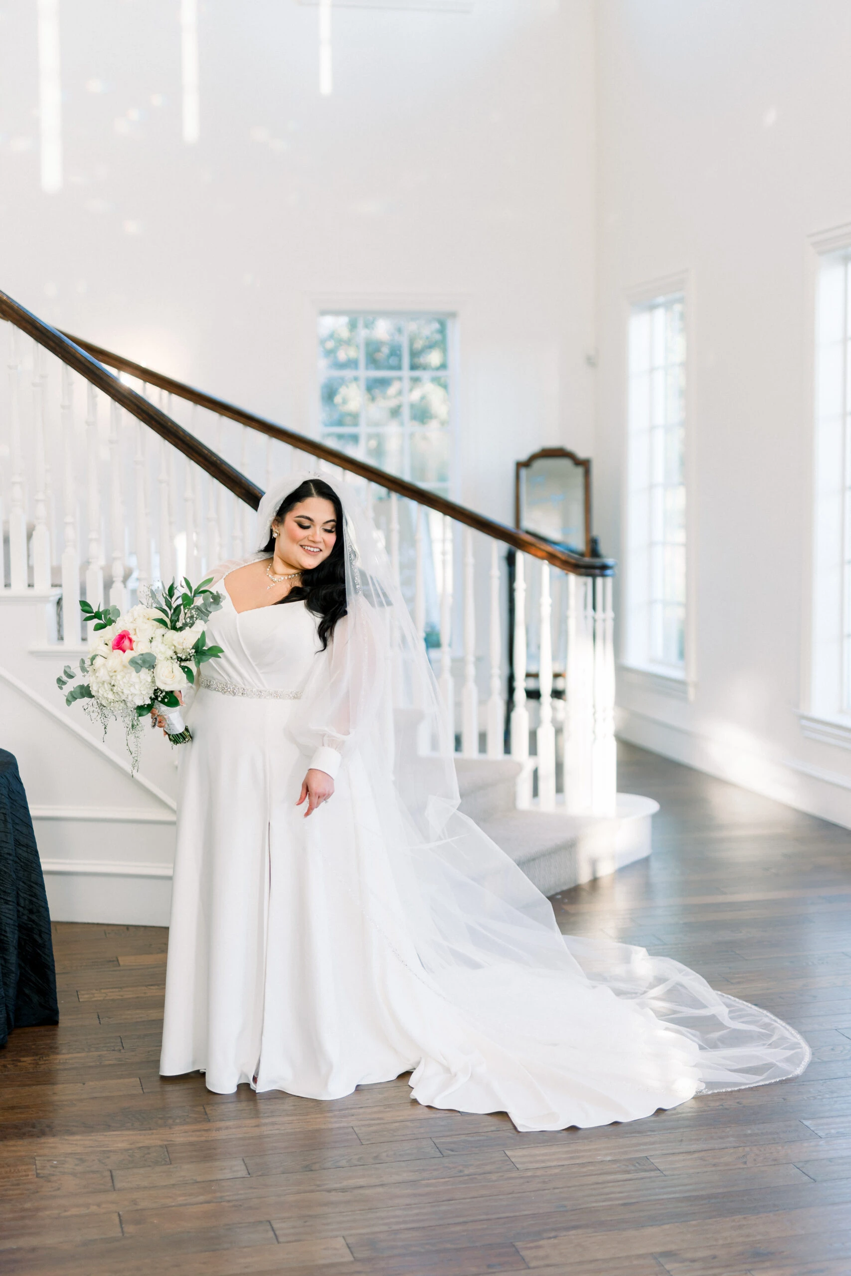 A bride in a white wedding gown poses inside next to a staircase, holding a bouquet and smiling.