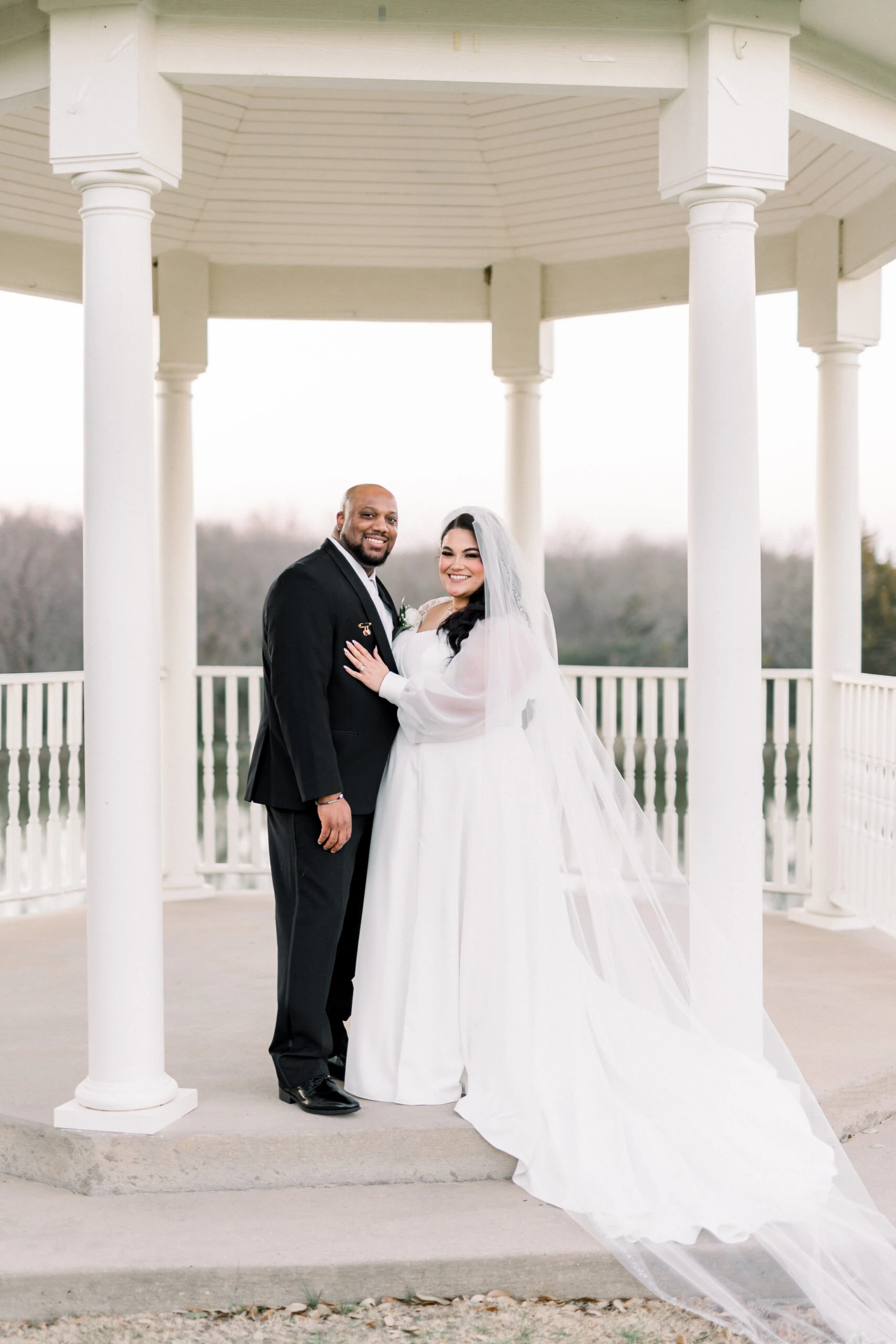 A couple stands under a gazebo, the bride in a flowing white gown with a long veil, and the groom in a black suit, both smiling.