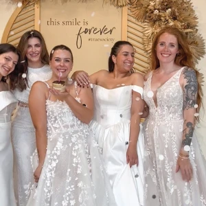 Five women dressed in elegant wedding gowns stand together, smiling joyfully. They are standing in front of a decorative backdrop with dried floral arrangements and a sign that reads 'this smile is forever #truesociety.' The women are celebrating, and one of them is holding a small dessert with a candle