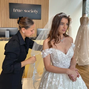 A bride wearing a lace off-the-shoulder wedding gown and a tiara stands as a woman, dressed in black, adjusts her veil. They are in a bridal boutique with a sign that reads 'true society' in the background. Mannequins displaying other wedding dresses are visible in the background