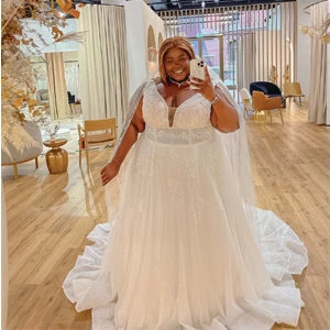 A bride wearing a sparkling lace wedding gown with a plunging neckline and a long veil smiles as she takes a selfie in a bridal boutique. The boutique has a warm and inviting atmosphere with modern decor, including chairs, tables, and floral arrangements